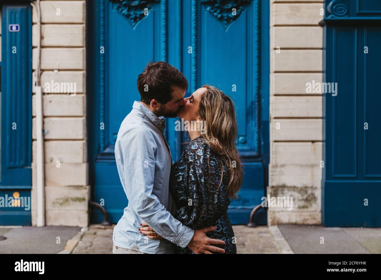 Side view of happy young couple in casual clothes hugging and kissing while standing against aged stone building with blue doors on city street Stock Photo