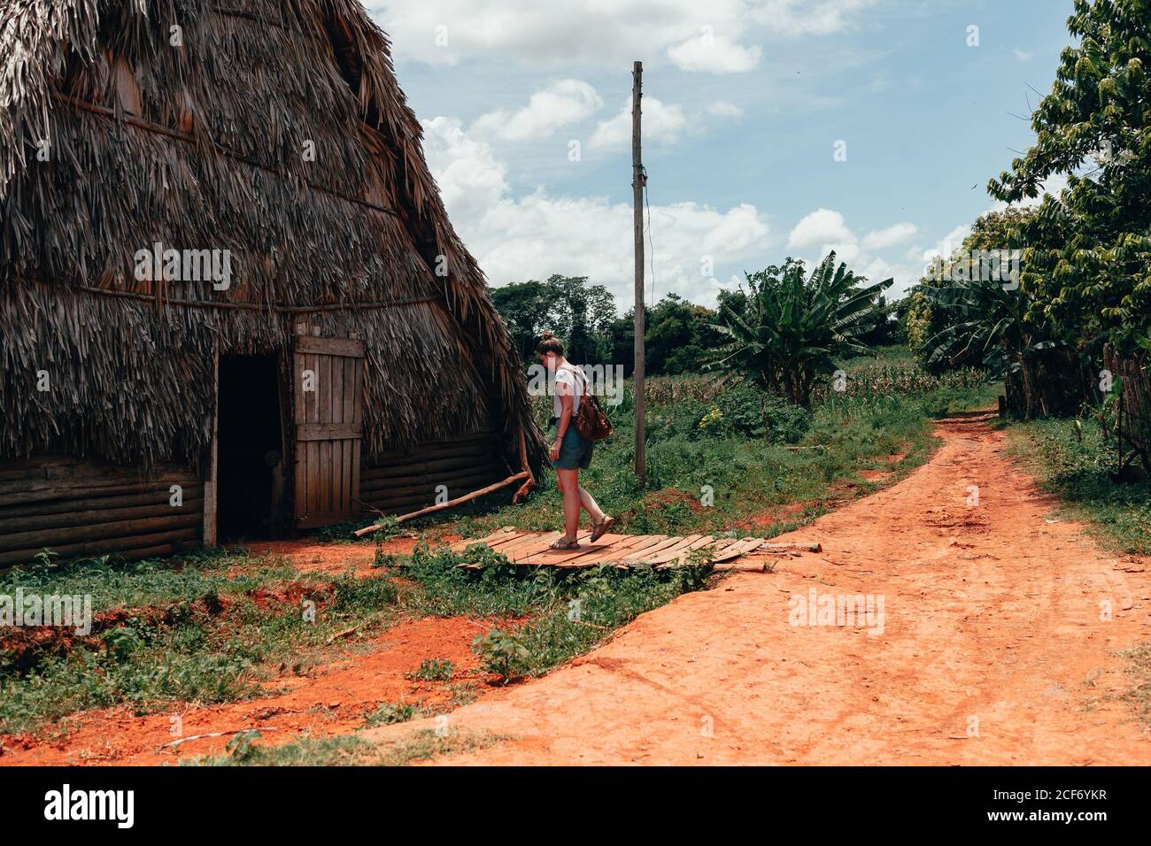 Resting lady in stylish casual clothing standing in doorway of big rural house with thatched roof and blue sky on background in Cuba Stock Photo