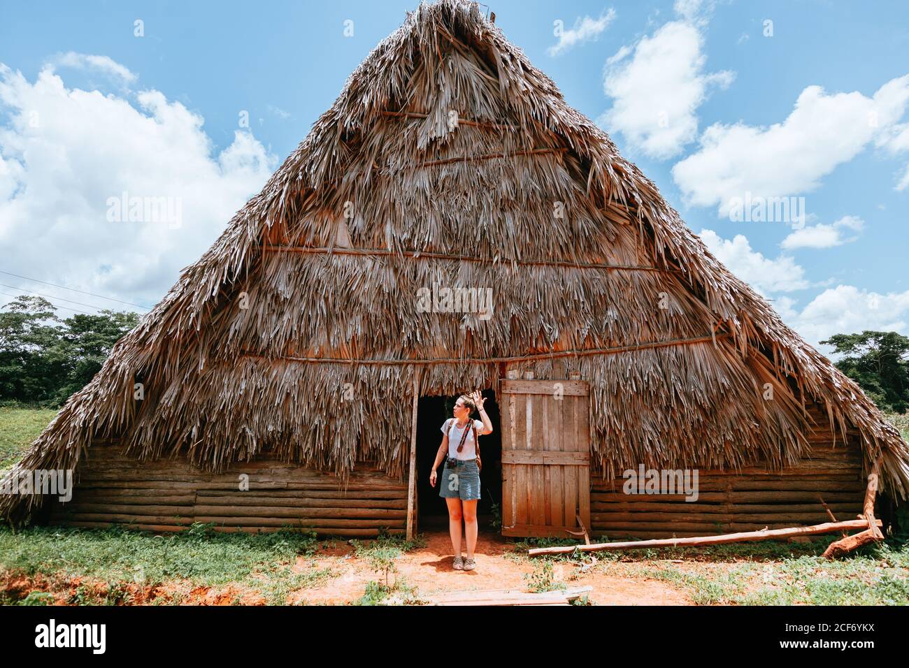 Resting lady in stylish casual clothing standing in doorway of big rural house with thatched roof and blue sky on background in Cuba Stock Photo