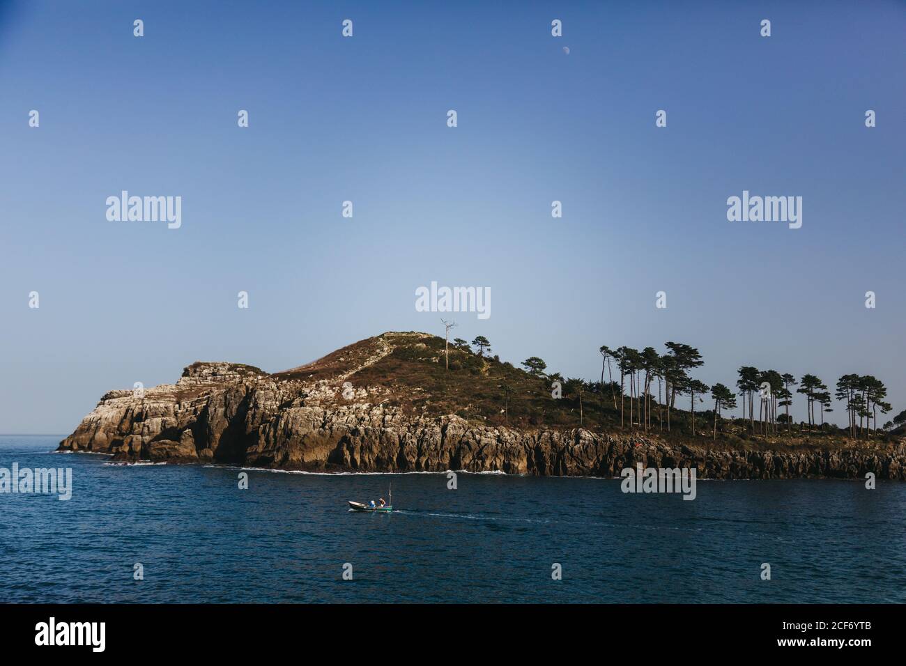 Lonely fishing boat moving on bay with tranquil water against rocky shore with tall evergreen trees at foot of hill under clear blue sky in Spain Stock Photo