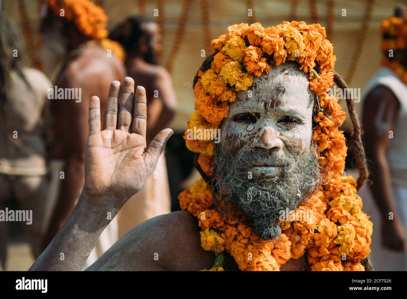 Allahabad city, India - FEBRUARY, 2018: Hindu sadhu with face painted with ash and saffron flowers around head and neck standing on street and waving hand at camera during Prayag Kumbh Mela Festival Stock Photo