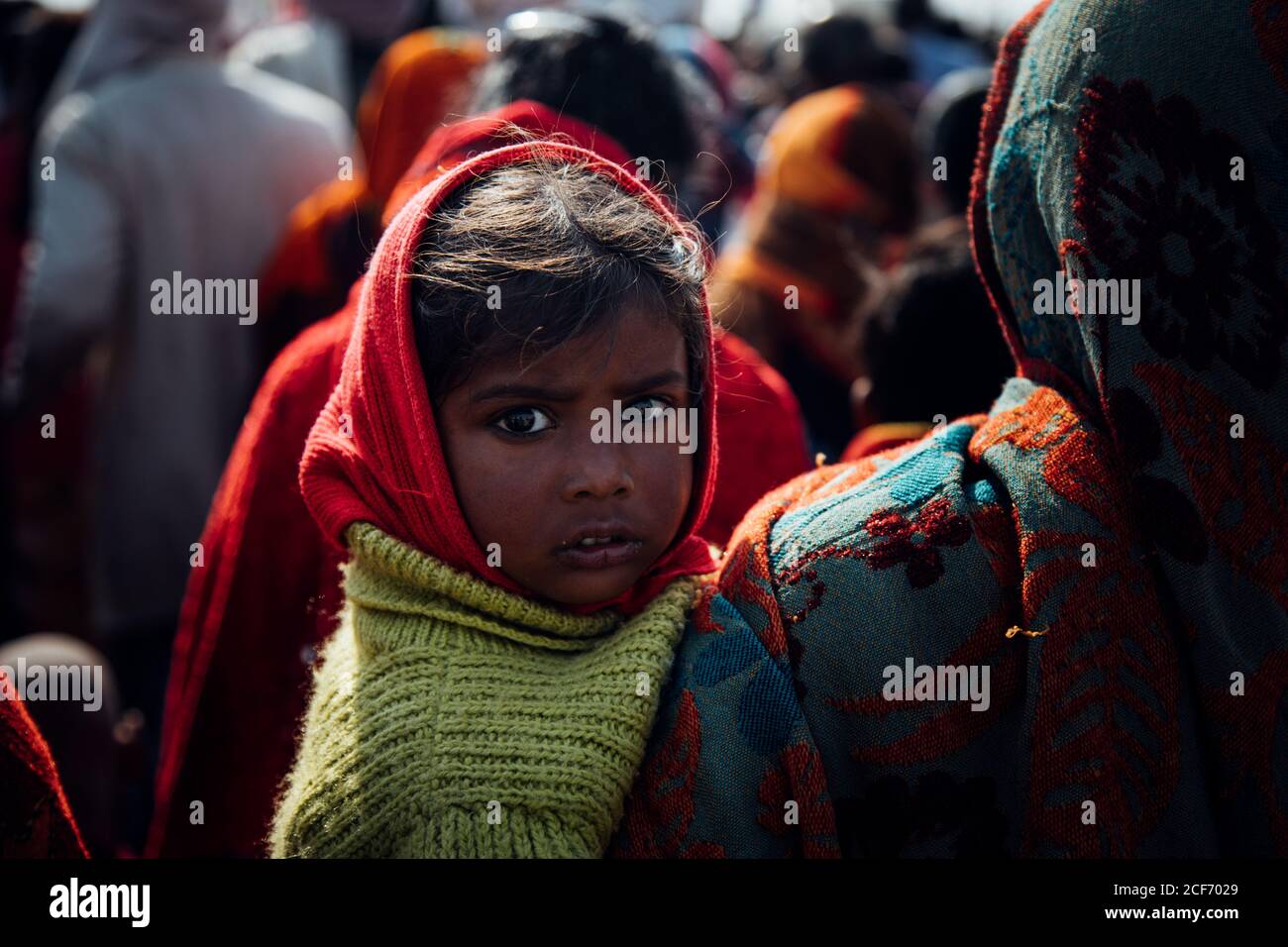 Allahabad city, India - FEBRUARY, 2018: Little girl in red headscarf and green knitted sweater sitting in arms of crop woman walking on street in crowd of pilgrims during Prayag Kumbh Mela Festival Stock Photo