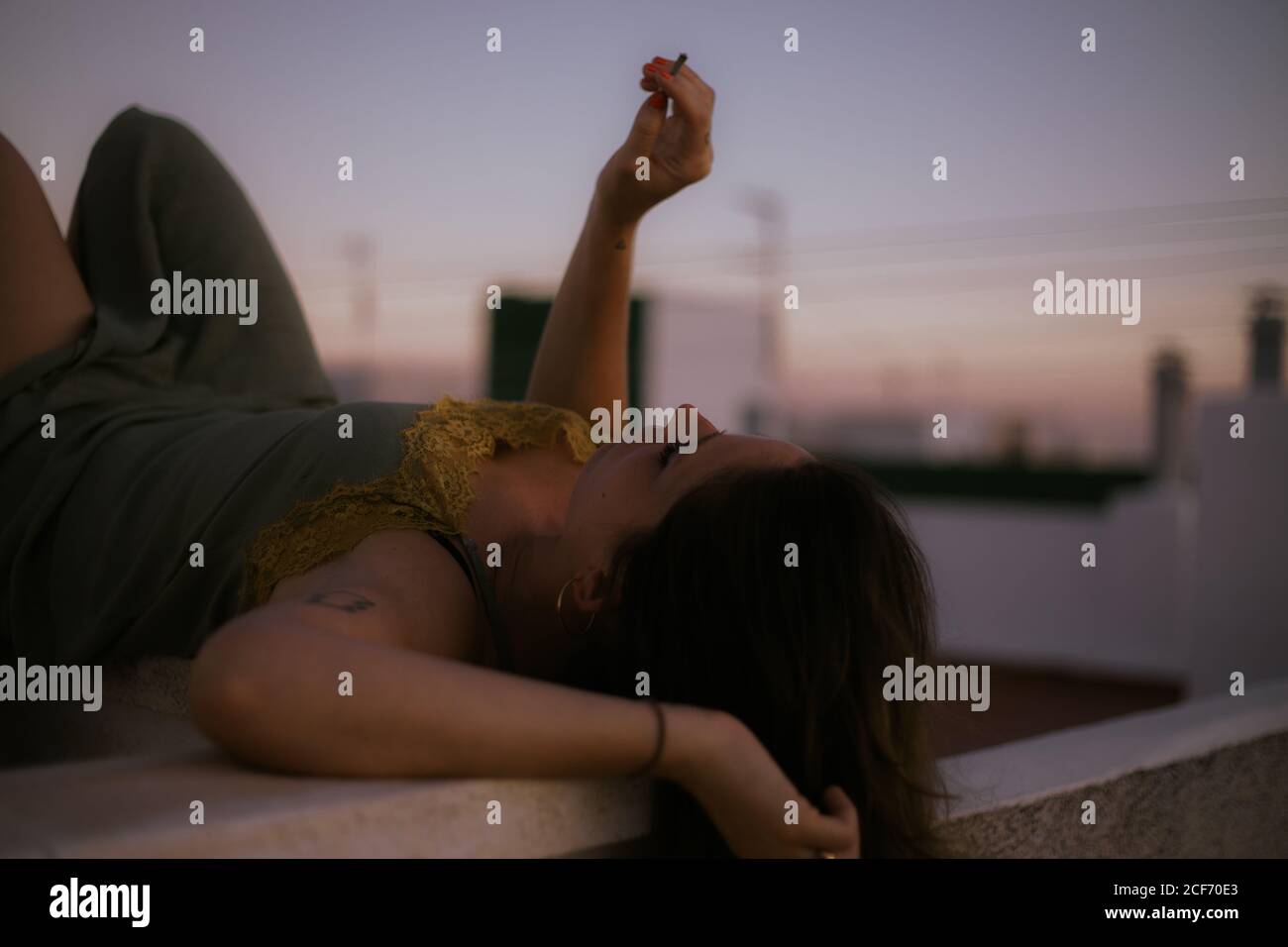 Side view of peaceful lady lying with closed eyes on fence of balcony and smoking cigarette with sunset on blurred background Stock Photo