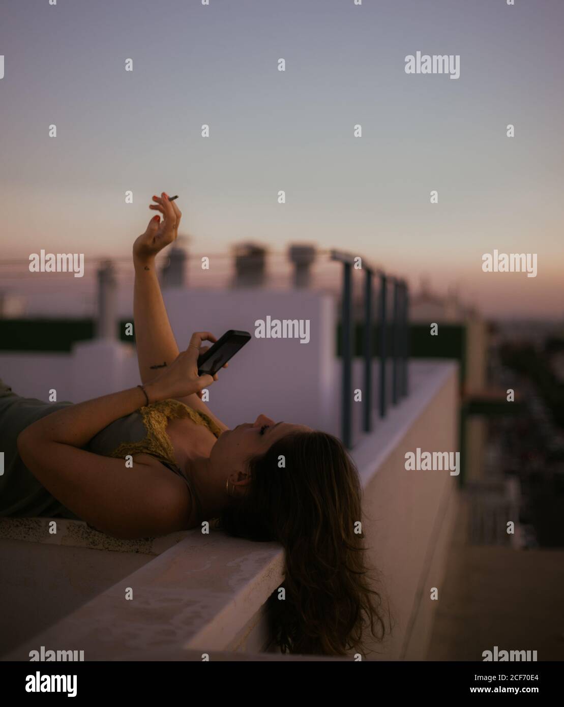 Side view of peaceful lady lying on fence of balcony smoking cigarette and using mobile phone to take a picture with sunset on blurred background Stock Photo