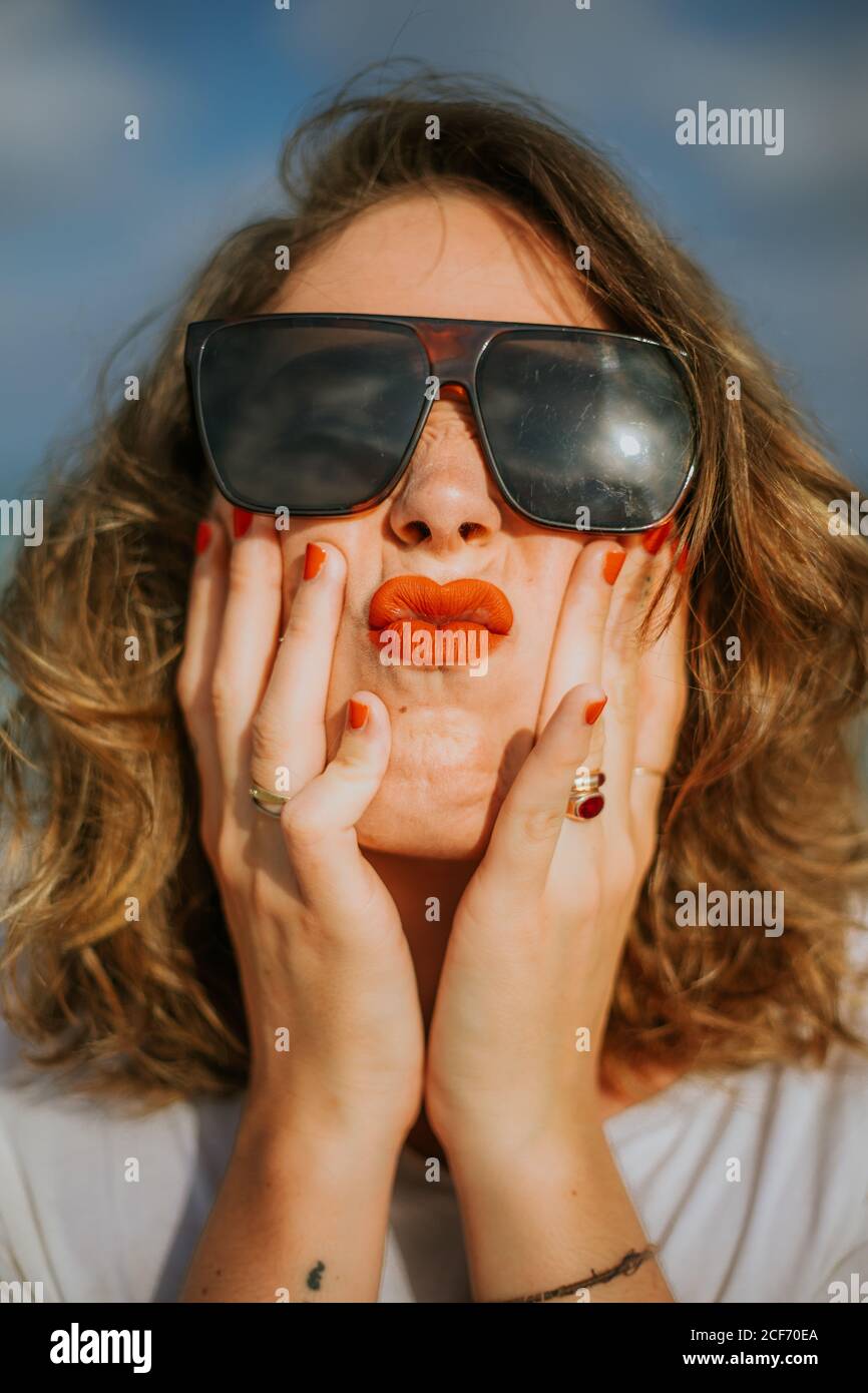 Stylish brown curly haired Woman with red lipstick in trendy sunglasses looking at camera while squeezing face in palms Stock Photo