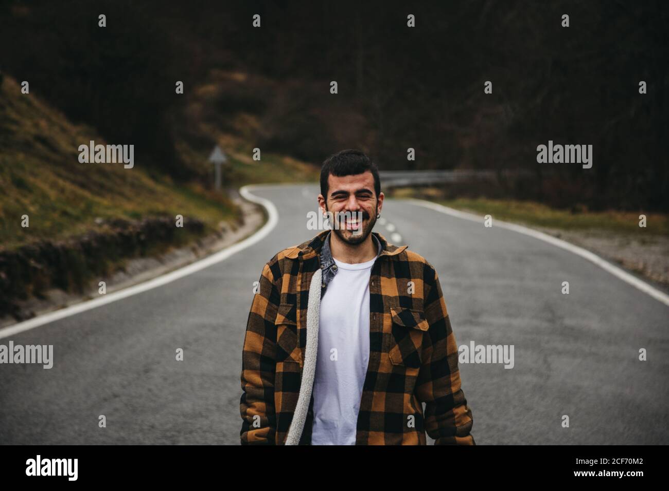 Bearded black haired cheerful man in casual wear smiling and looking at camera while standing on empty road in autumn countryside Stock Photo