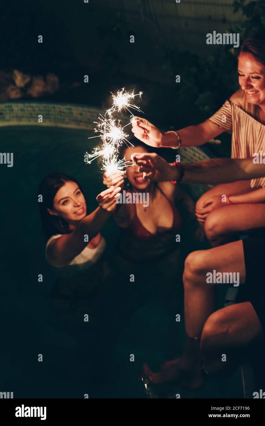 Group of young people smiling and burning sparklers while having party near pool at night Stock Photo