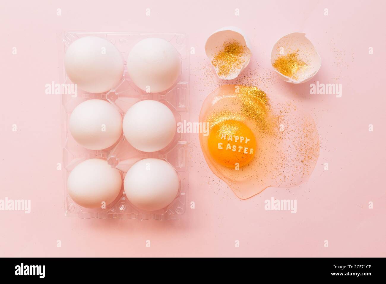 Top view of broken glittered egg with text Happy Easter and raw eggs in plastic box on pink background Stock Photo