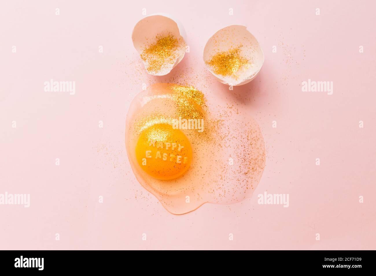 From above raw broken egg with text Happy Easter on yellow yolk covered in glitter and broken eggshells on pink background Stock Photo