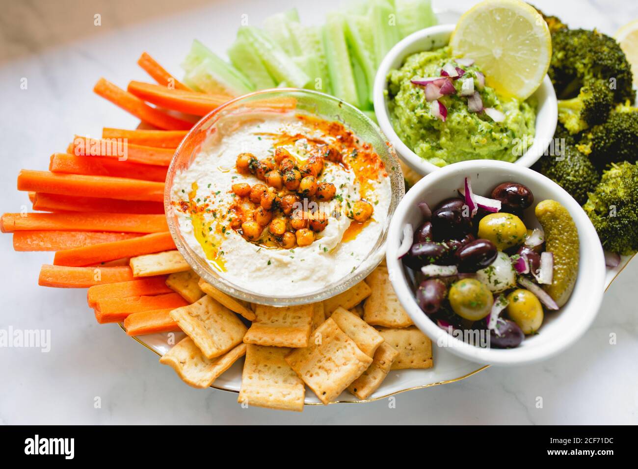 From above appetizing healthy hummus green sauce cut carrot cucumbers and green broccoli salted cucumbers decorated with sliced lemon in white bowls on table Stock Photo