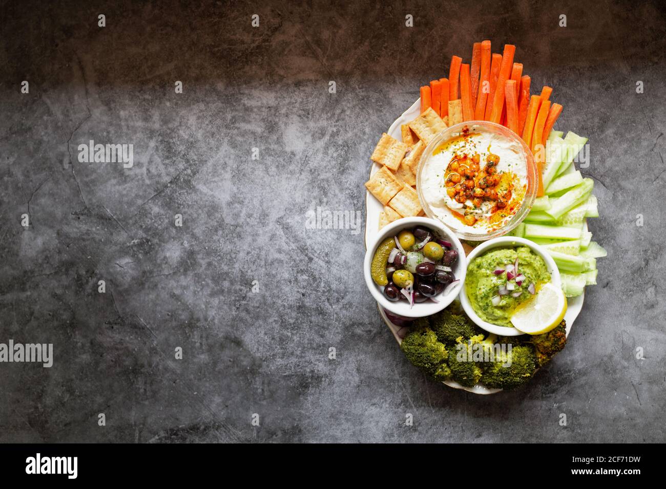 From above appetizing healthy hummus green sauce cut carrot cucumbers and green broccoli salted cucumbers decorated with sliced lemon in white bowls on table Stock Photo