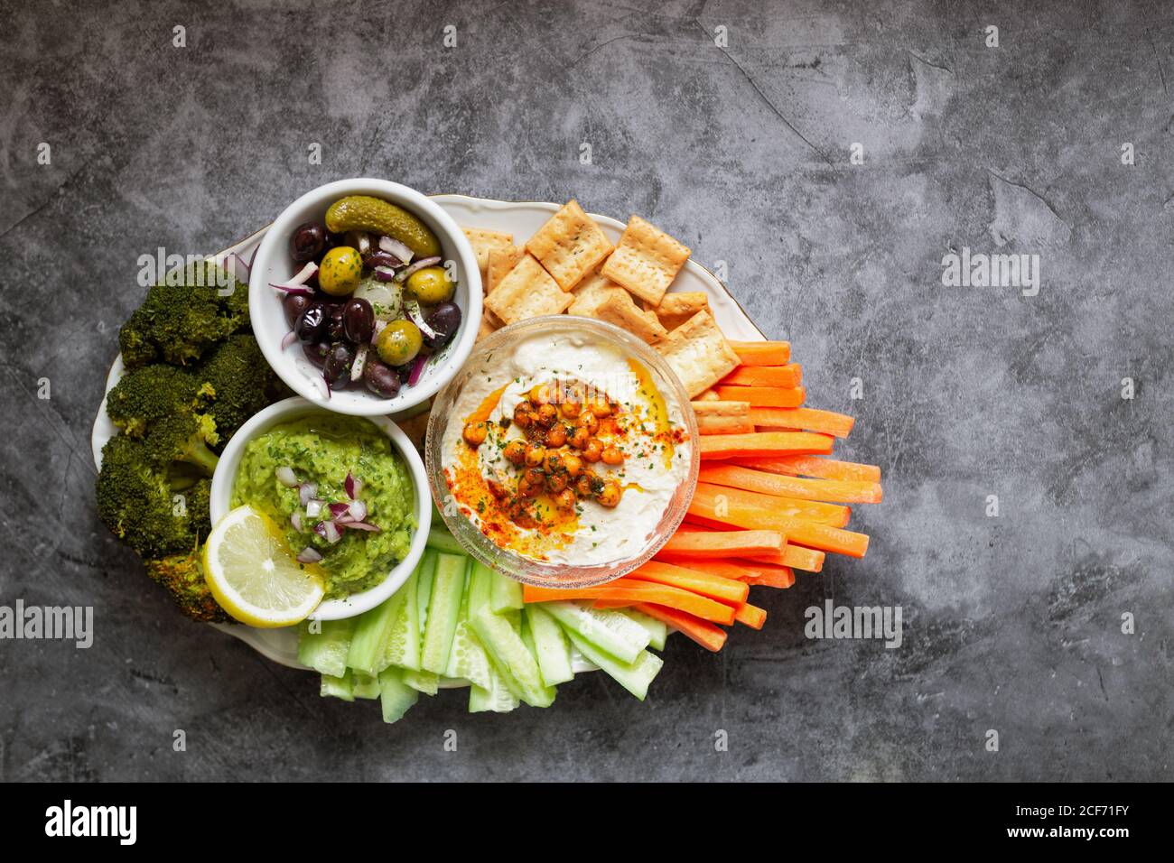 From above appetizing healthy hummus green sauce cut carrot cucumbers and green broccoli salted cucumbers decorated with sliced lemon in white bowls on table Stock Photo