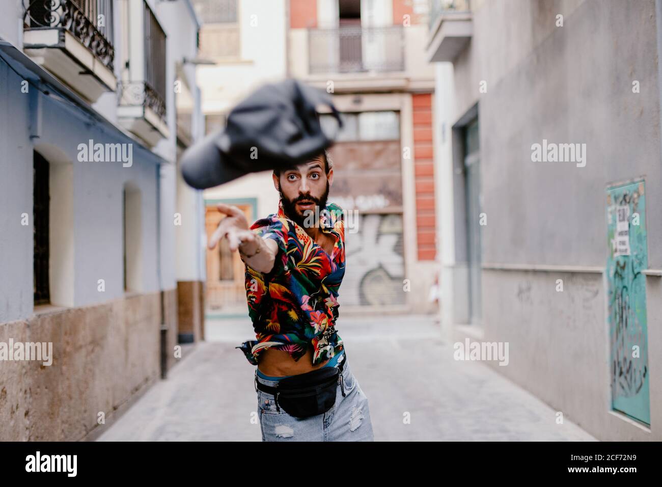 Concentrated young bearded man standing on city alley and throwing cap at camera Stock Photo