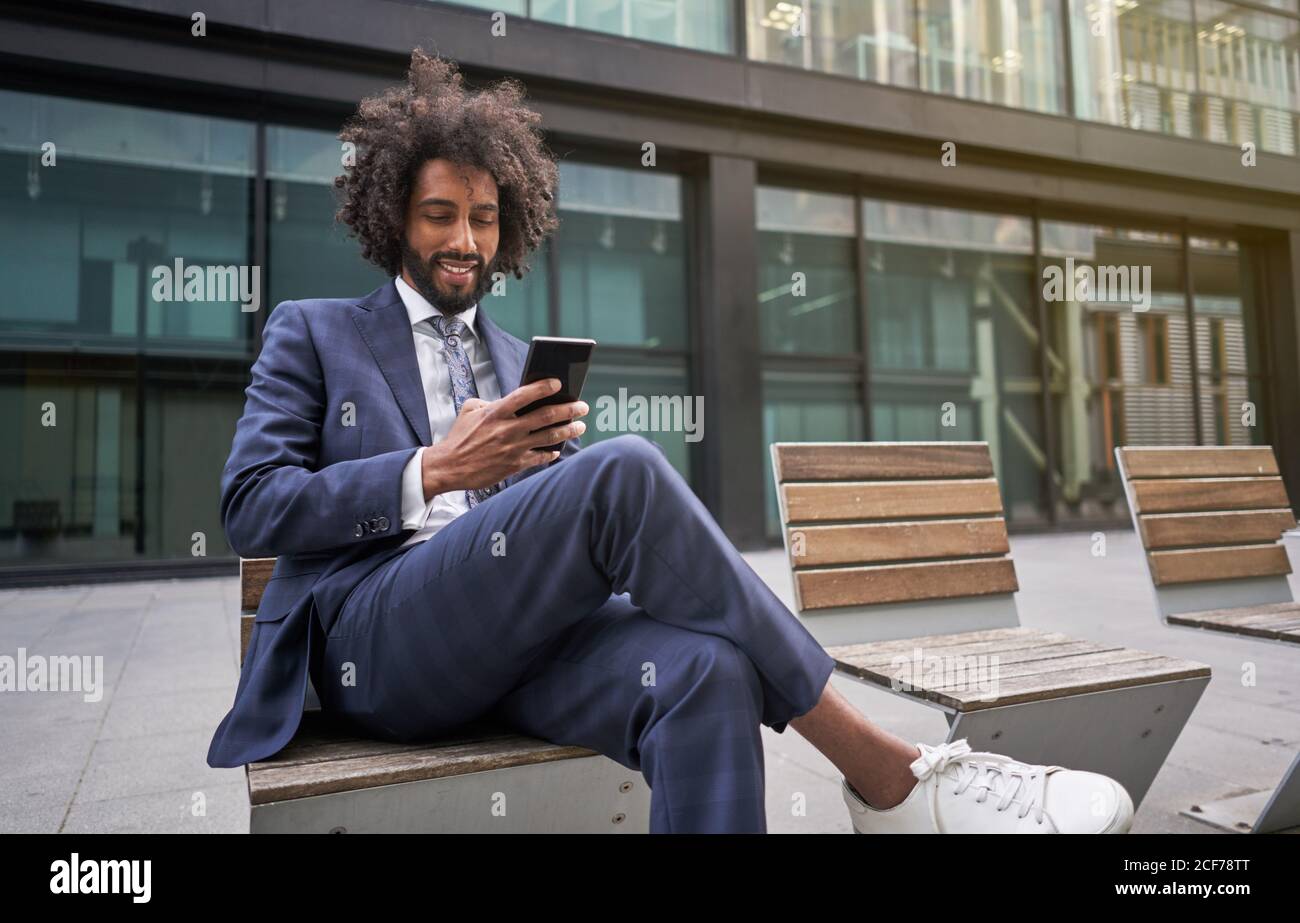 Joyful ethnic man in suit sitting outside and messaging Stock Photo