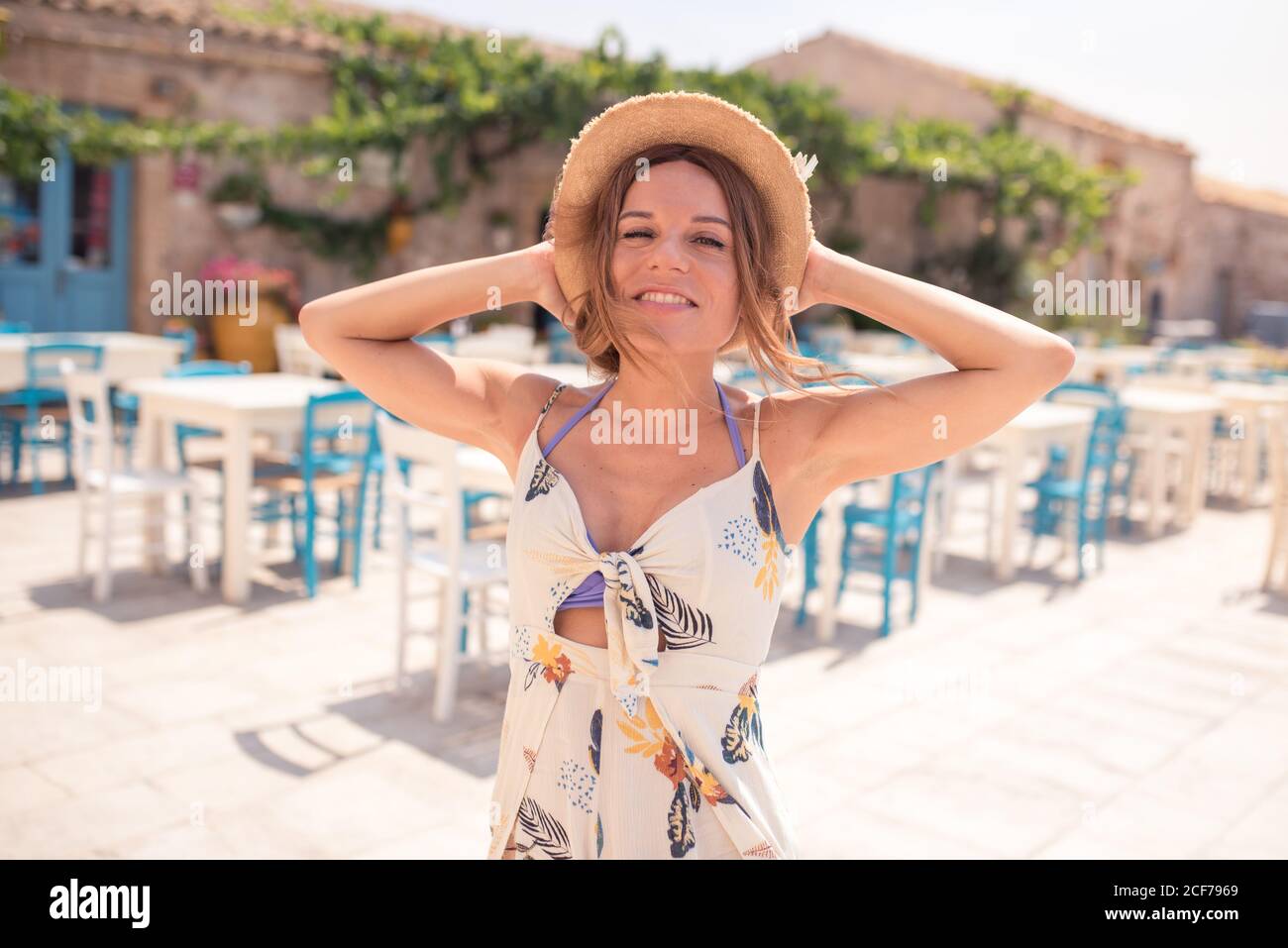 Alluring adult female in straw hat wearing light dress looking at camera while standing alone on cafe terrace against blue and white furniture in sunlight Stock Photo