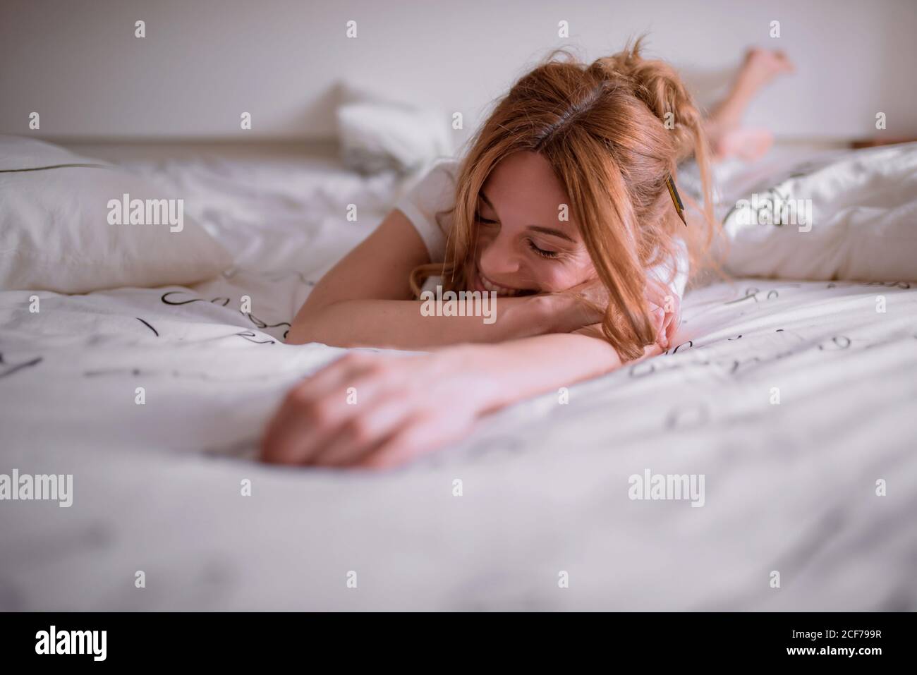 Young satisfied female with pencil in red hair having rest after awakening at weekend morning and home lying on stomach on cozy bed with white linen and smiling with closed eyes Stock Photo