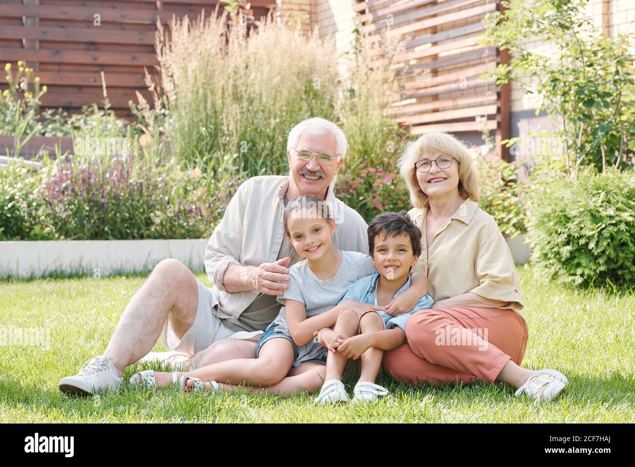Group portrait of cheerful grandma, grandpa and two grandchildren sitting together on lawn in backyard looking at camera smiling Stock Photo