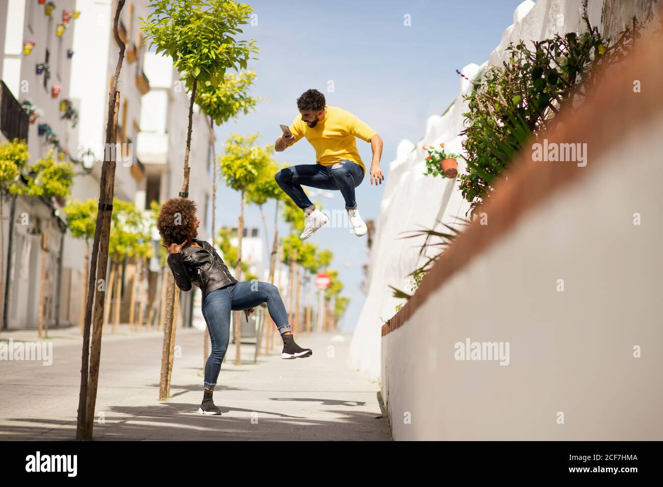 Stylish young guy leaping up and taking picture of funny female while spending time on city street together Stock Photo