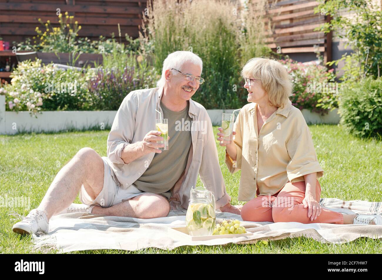 Modern married seniors sitting on lawn in backyard drinking fresh homemade lemonade and talking about something Stock Photo