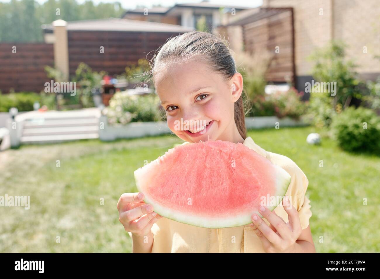 Medium close-up portrait shot of cheerful little girl standing in backyard holding piece of watermelon smiling at camera Stock Photo