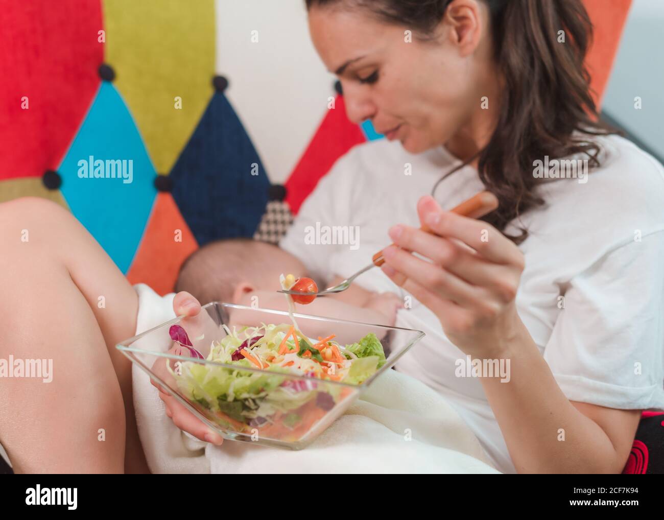 Side view of black haired young mother in casual wear sitting on armchair and breastfeeding baby while eating healthy salad Stock Photo
