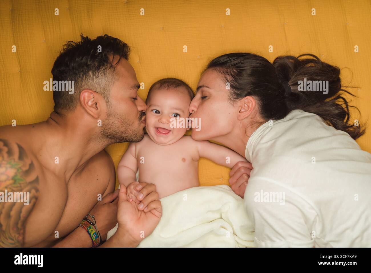 Top view of young happy parents kissing in cheeks little satisfied baby while lying on bed Stock Photo