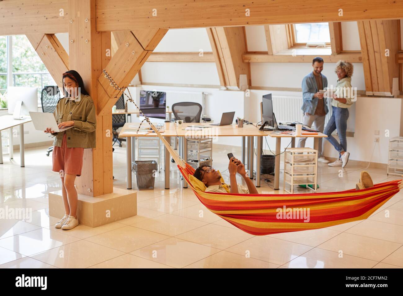 Young man relaxing in hammock and using his mobile phone with his colleagues using digital tablets they have a time out at modern office Stock Photo