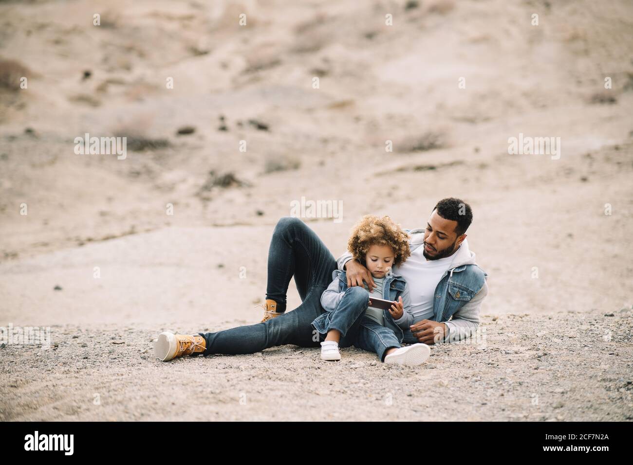 African American bearded casual man lying on sandy ground leaning on elbow and bending leg at knee looking at curly ethnic toddler in denim clothes sitting beside with mobile phone Stock Photo