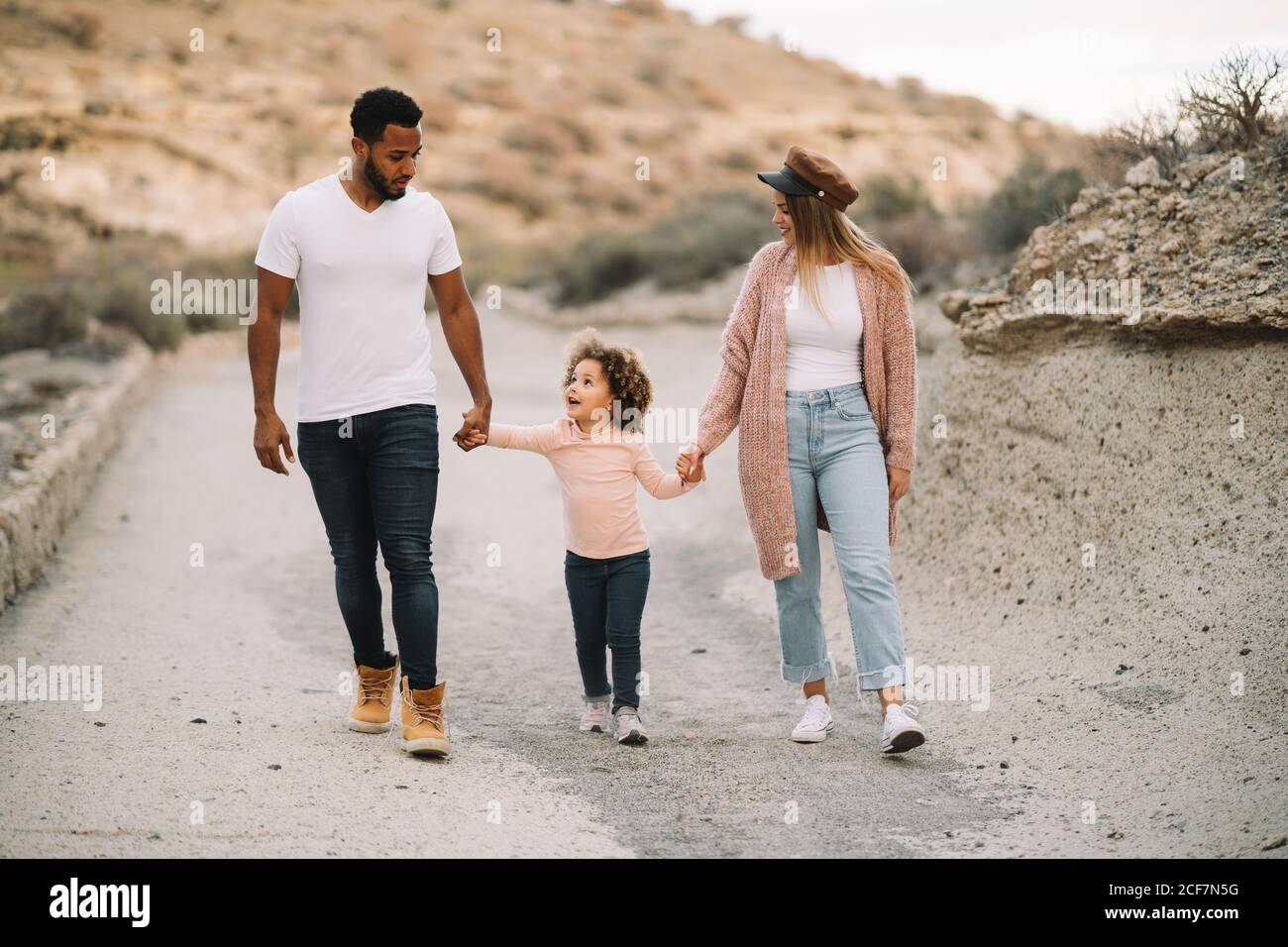Happy diverse mother father and curly child wearing bright casual clothes strolling on nature holding hands at daytime Stock Photo
