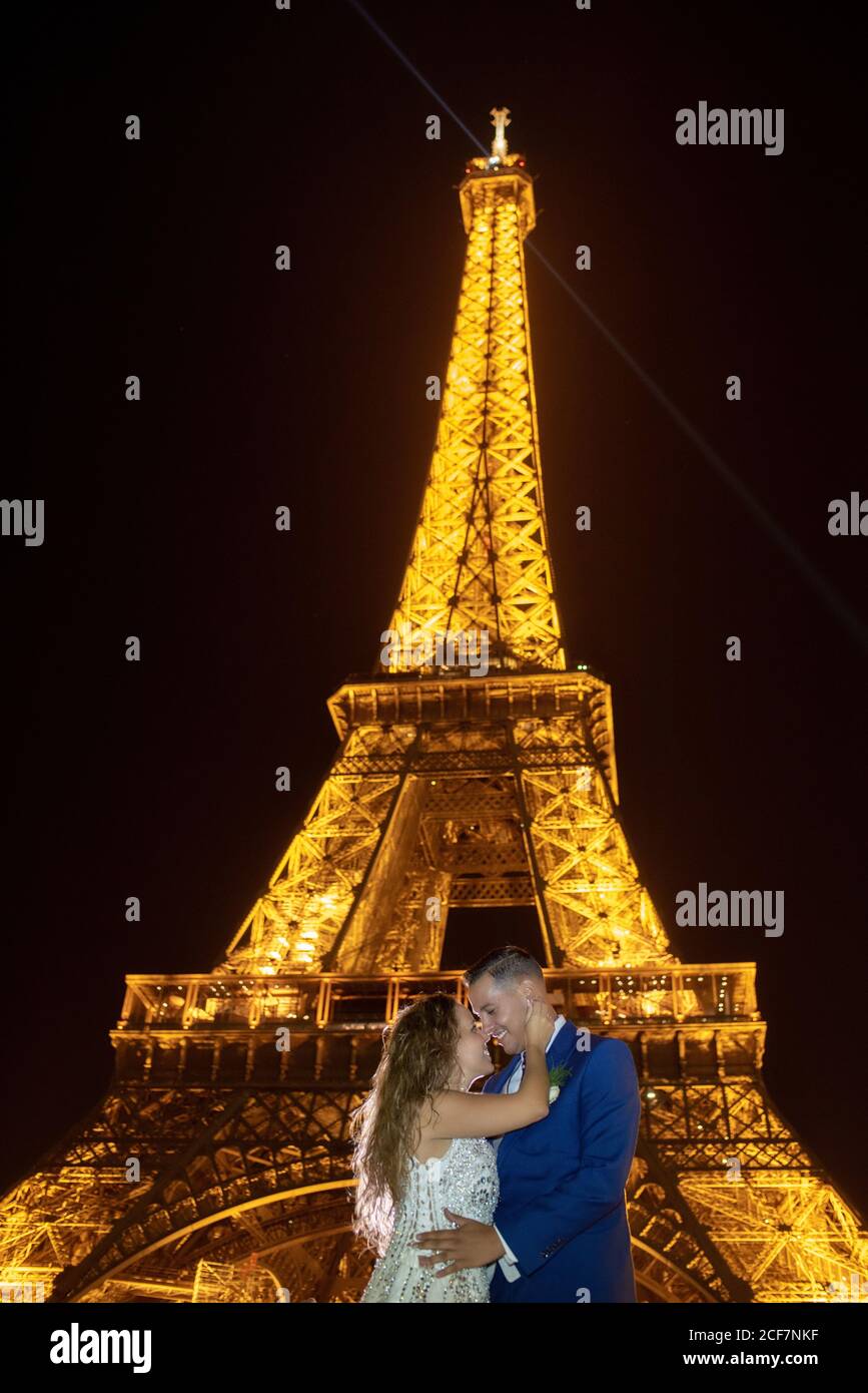 Joyful groom in blue suit and bride in white wedding dress embracing while  smiling and kissing on evening with Eiffel Tower on background at Paris  Stock Photo - Alamy