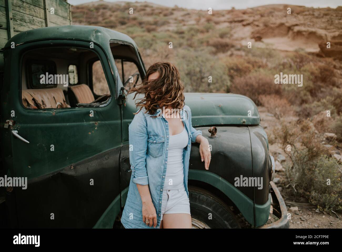 Young Woman in shorts and denim shirt standing by green truck with wooden body and looking away on nature background Stock Photo