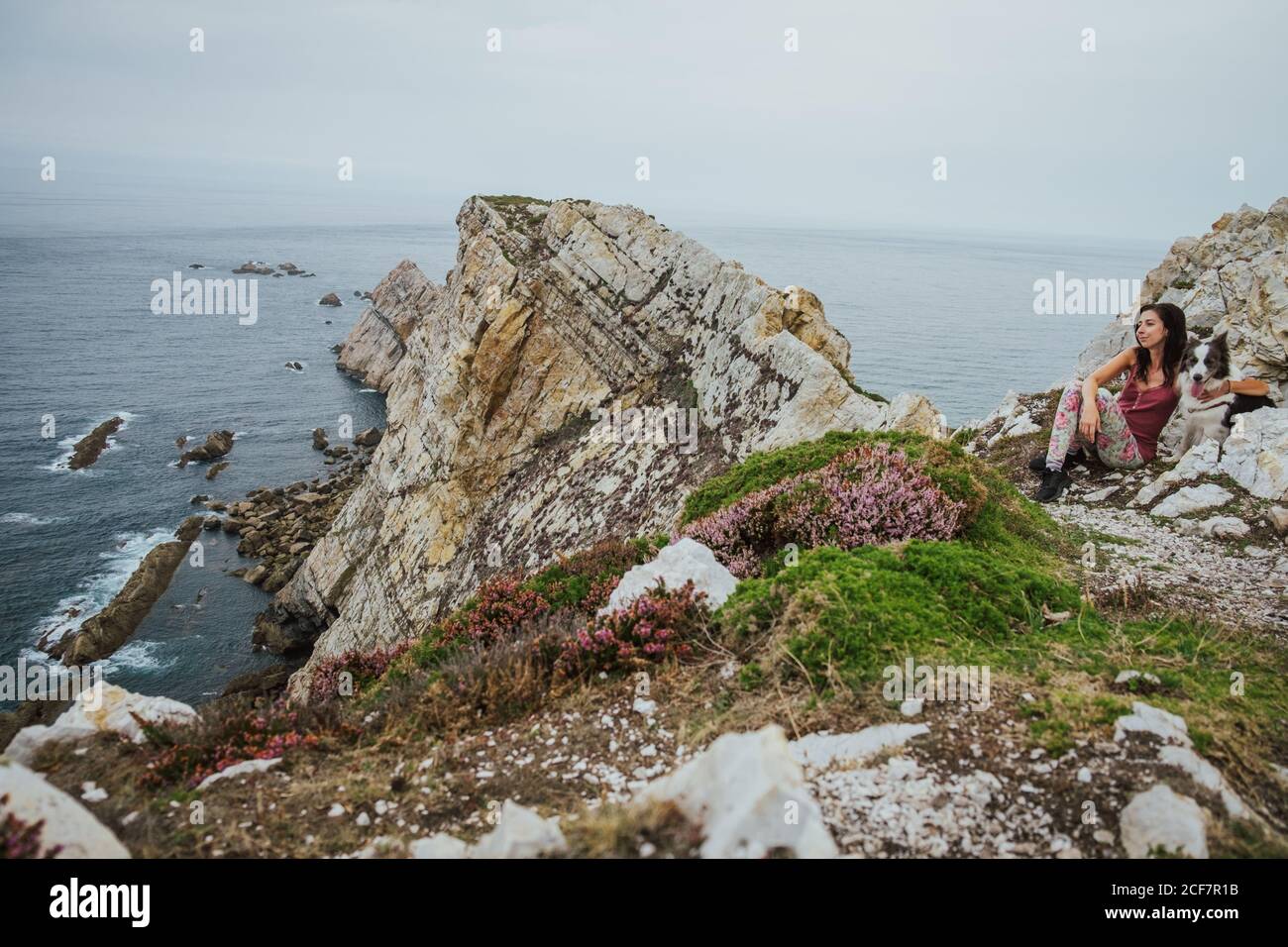Adult Woman in casual clothes looking away and enjoying views while sitting and hugging Border Collie dog on rocky shore against gray waves under cloudy sky in Asturias in summer Stock Photo