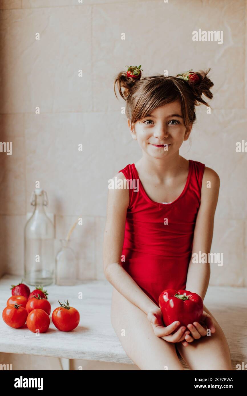 Cute barefoot girl in red bodysuit and with strawberries in her hair holding red pepper looking at camera sitting on counter near tomatoes in kitchen Stock Photo