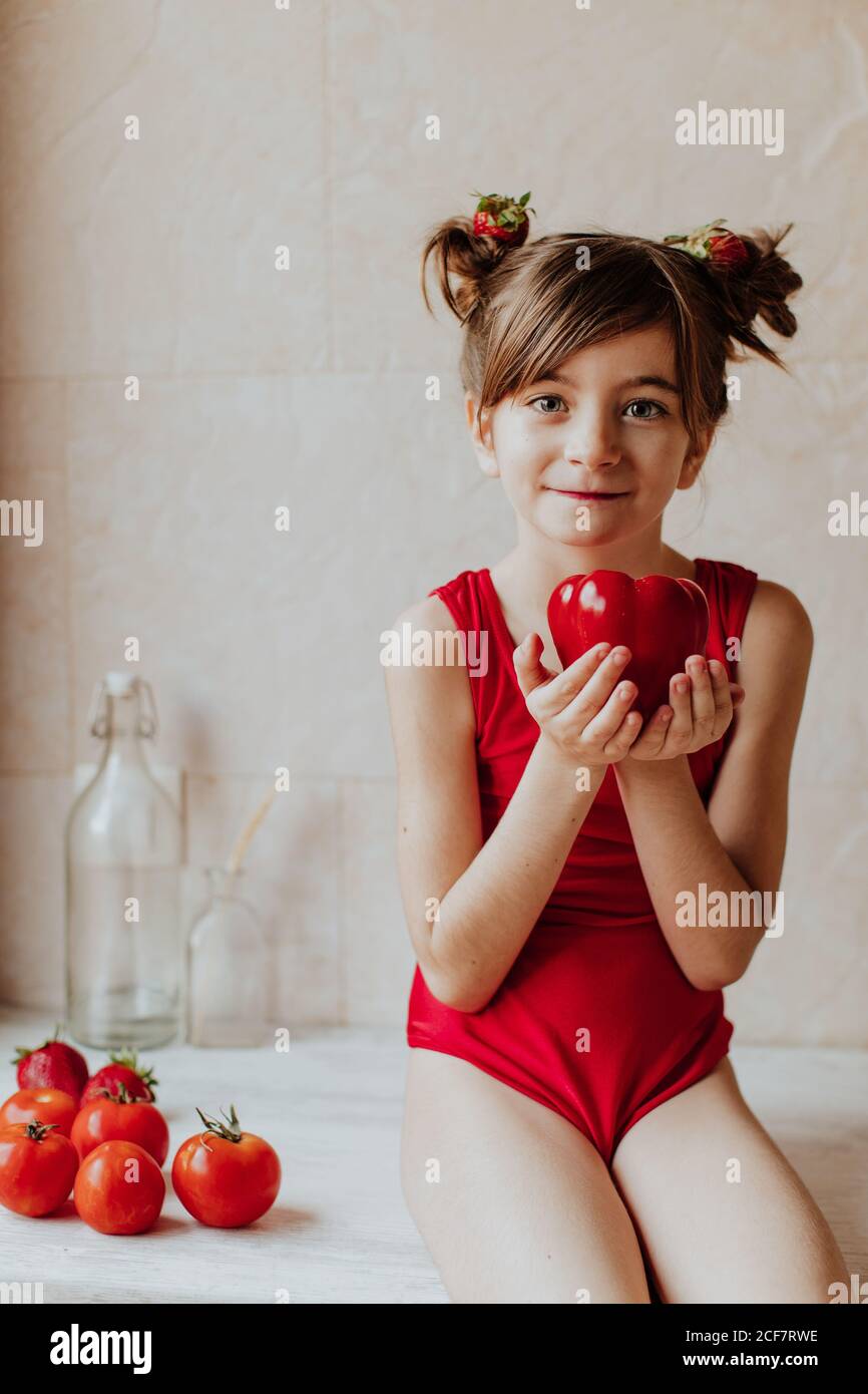 Cute barefoot girl in red bodysuit and with strawberries in her hair holding red pepper looking at camera sitting on counter near tomatoes in kitchen Stock Photo