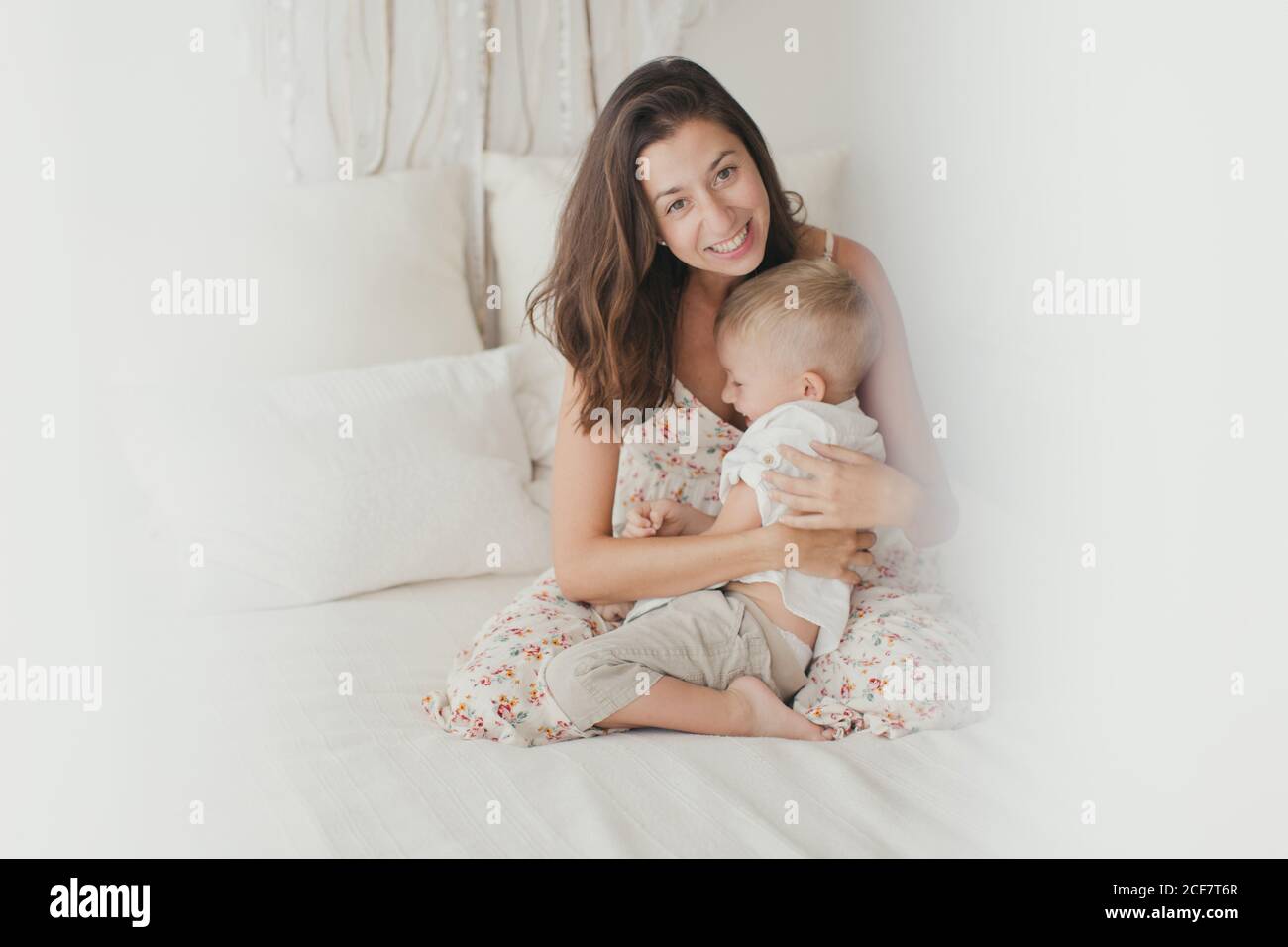 Satisfied brunette in white gown having fun with happy male toddler while embracing on bed Stock Photo