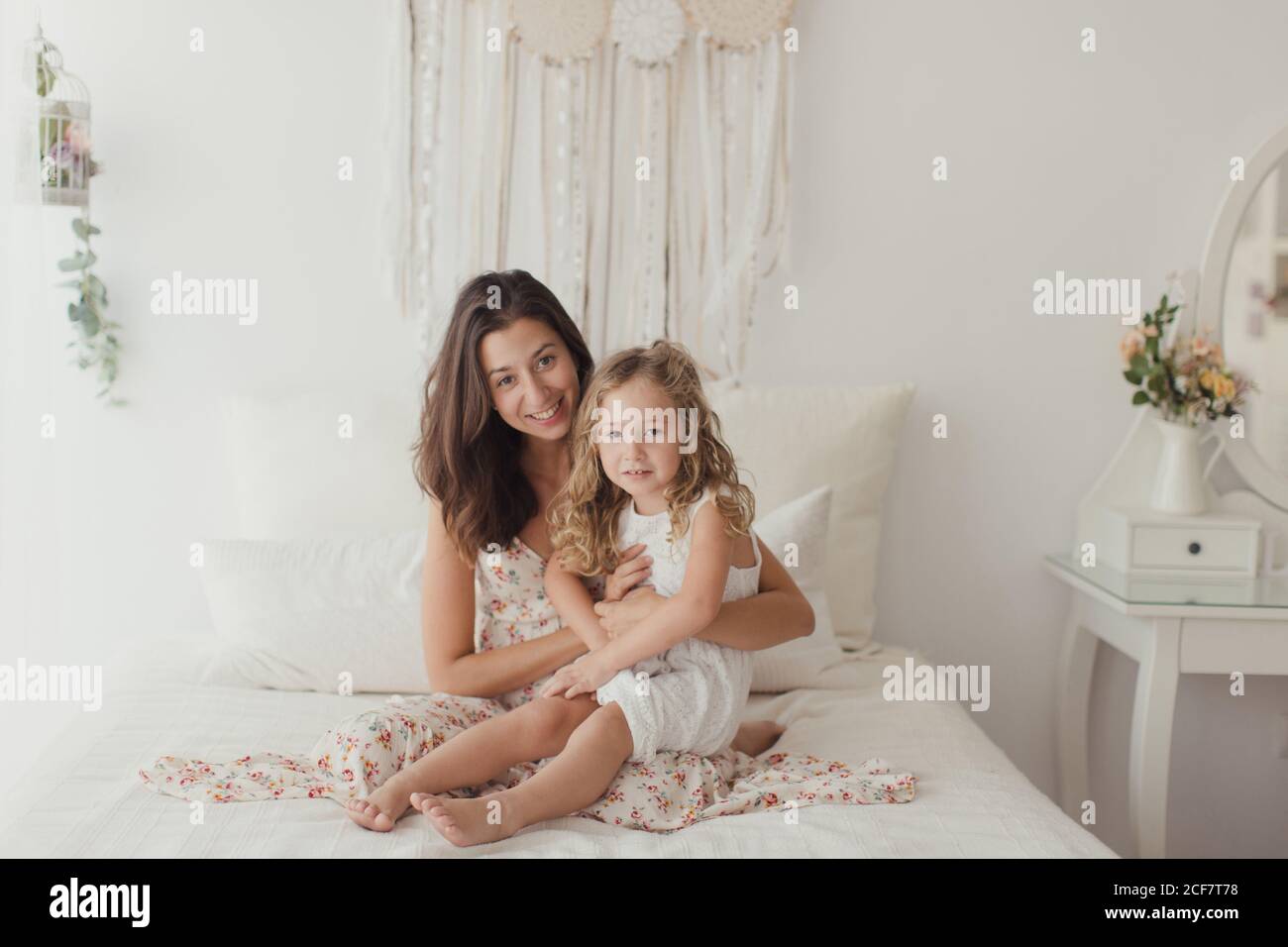 Satisfied brunette in white gown having fun with happy daughter while embracing on bed Stock Photo