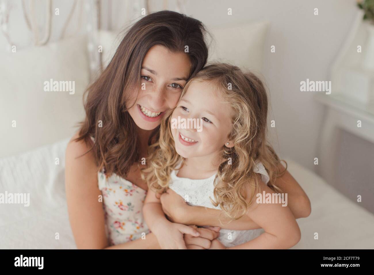 Satisfied brunette in white gown having fun with happy daughter while embracing on bed Stock Photo
