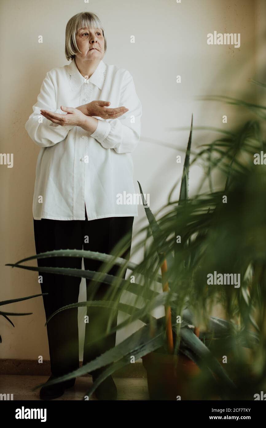 Focused female pensioner in white blouse and black trousers standing with crossed arms at wall looking away Stock Photo