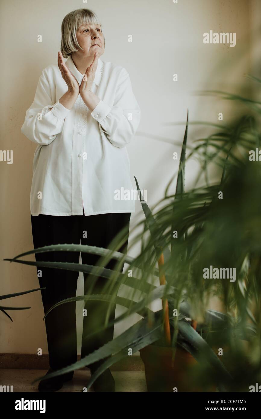 Focused female pensioner in white blouse and black trousers standing at wall making gestures with hands and looking away Stock Photo
