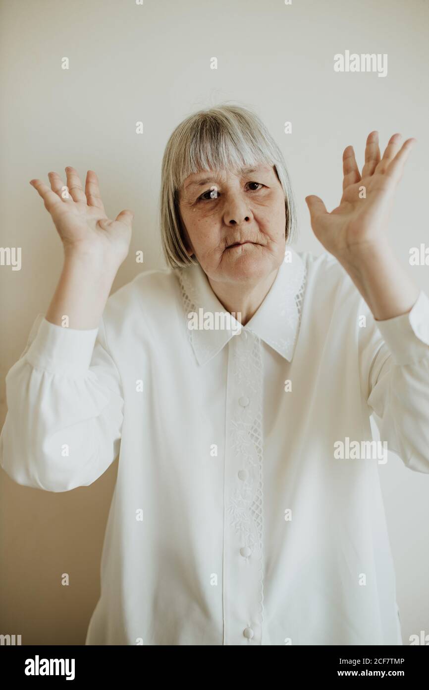 Sad elderly Woman in white blouse showing disagreement while rising hands up on light background looking at camera Stock Photo