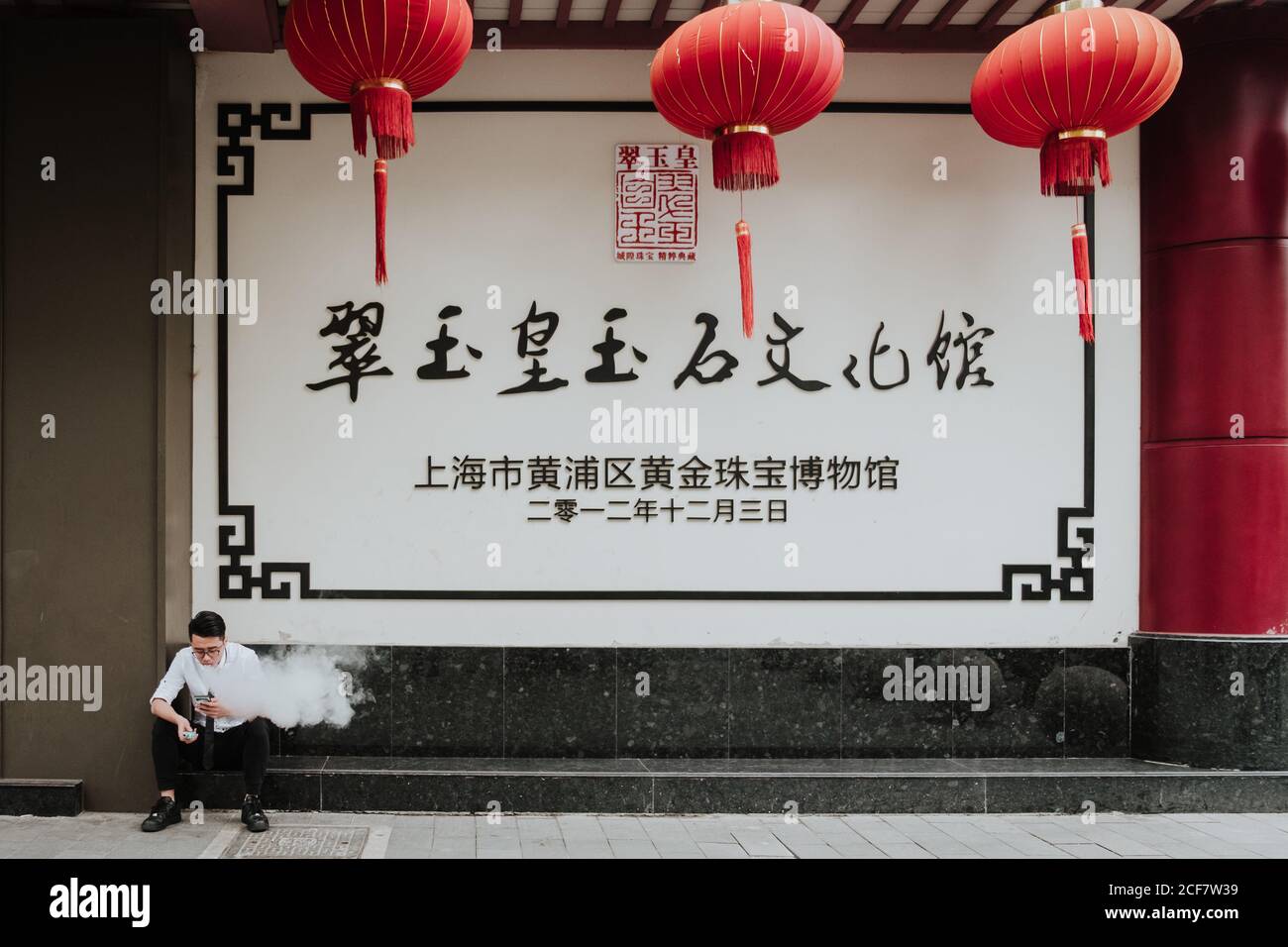 Shanghai, China - 12 December, 2018: Young man in formal wear smoking and using mobile phone while sitting next to  building with big signboard and red Chinese lanterns Stock Photo