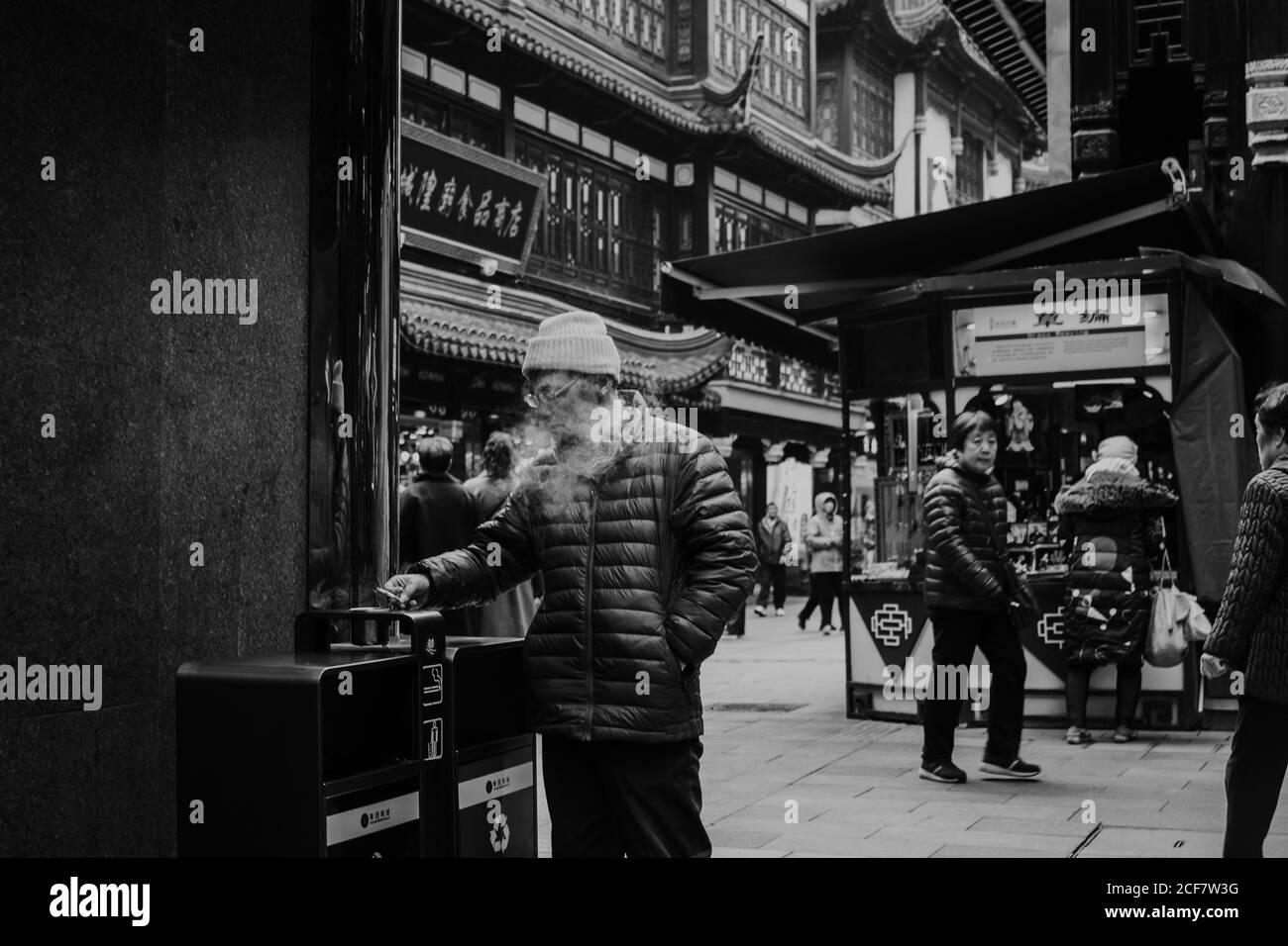 Shanghai, China - 12 December, 2018: Smoking adult male in warm jacket and knitted cap throwing cigarette to trash can while standing on street of Shanghai with traditional buildings and stalls Stock Photo