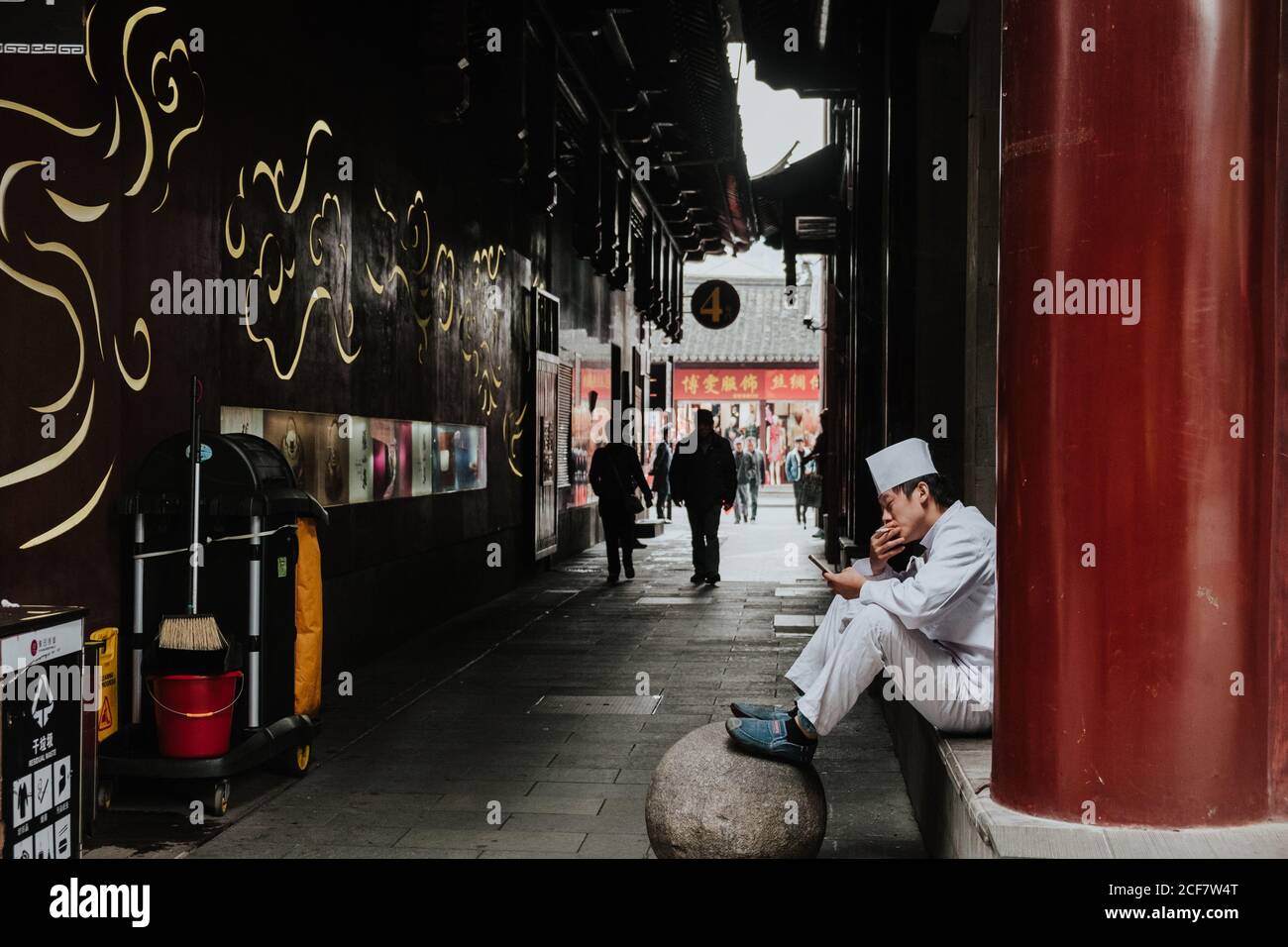 Shanghai, China - 12 December, 2018: Side view of young male in white uniform smoking and using smartphone while sitting next to building on narrow pedestrian street Stock Photo