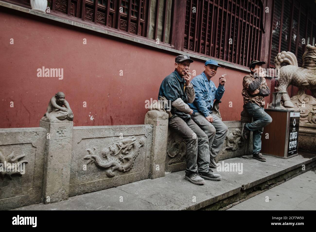Shanghai, China - 12 December, 2018: Group of adult Chinese male workers having break and smoking while sitting on stone fence next to building with red wall Stock Photo