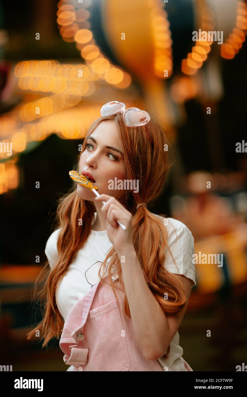 Charming red haired female teenager with ponytails and heart shaped eyeglasses on head standing in amusement park with illuminated attractions and eating lollipop Stock Photo
