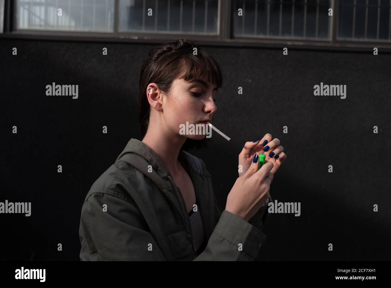 Side view of rebel young brunette in stylish casual jacket with blue nails lighting up cigarette at city street Stock Photo