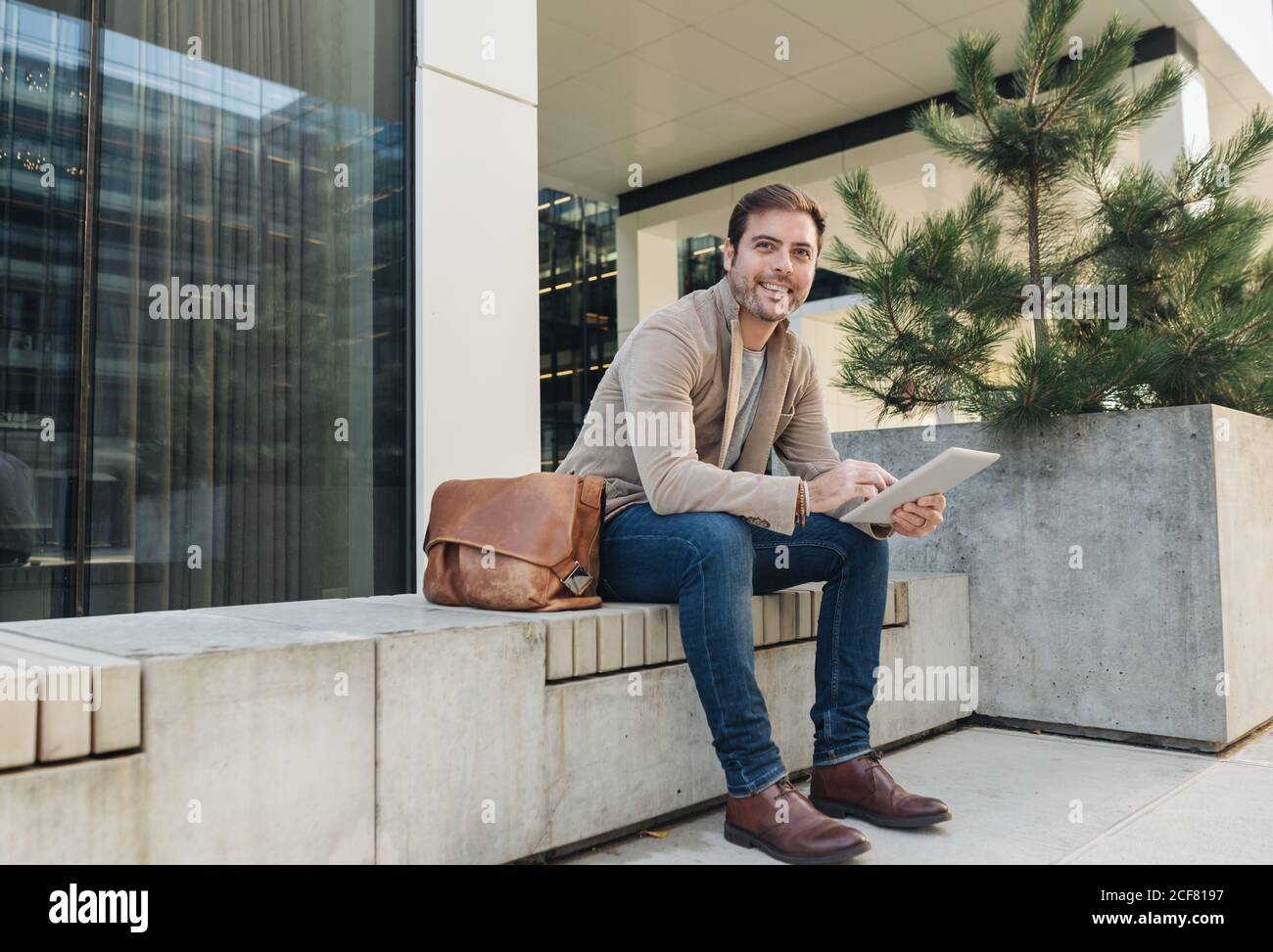 man manager smiling and browsing on a tablet while sitting outside contemporary building on city street Stock Photo