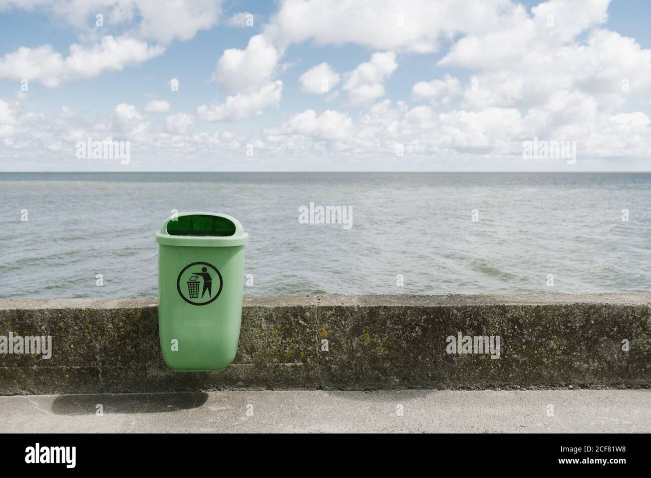 Green litter bin on clean empty concrete sidewalk parapet of seaside on cloudy day Stock Photo