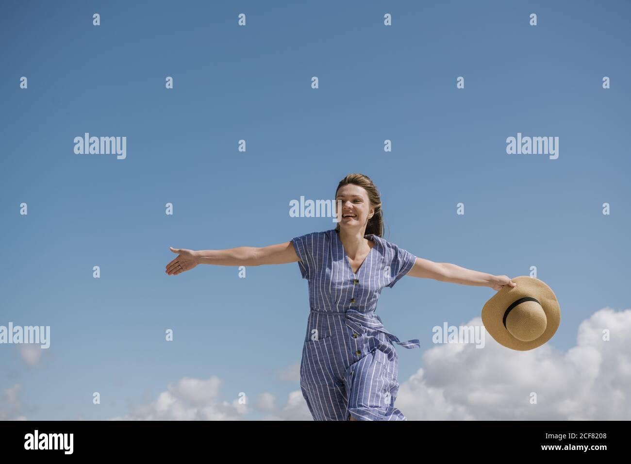 Content adult Woman with blowing hair and in sundress walking with straw hat in hand on cloudy sky background Stock Photo