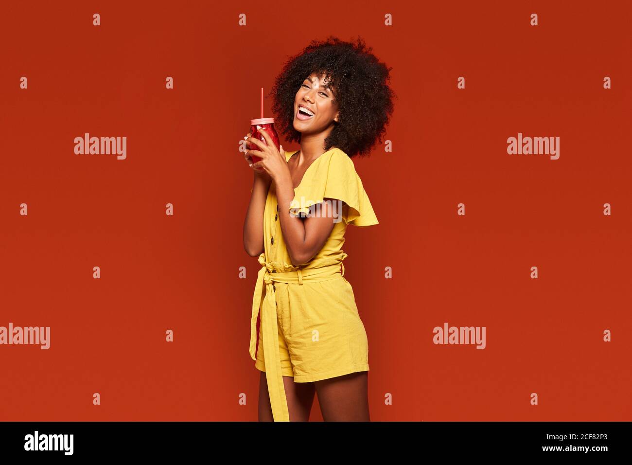 Cheerful young African American lady with curly hair holding red jar with straw and enjoying beverage on red background looking at camera Stock Photo